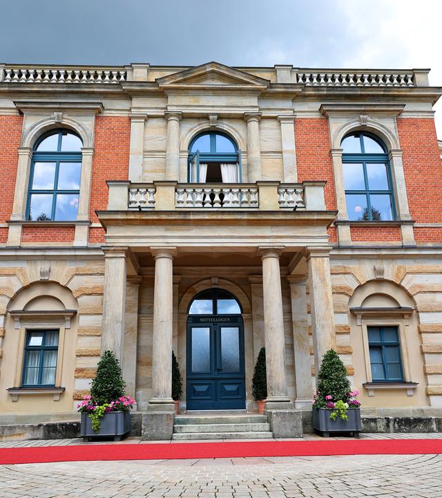 Ein großes zweistöckiges historisches Gebäude aus roten Backsteinen und sandfarbenem Stein mit Säuleneingang und Balkon im oberen Stockwerk.