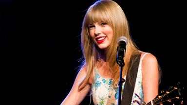 Taylor Swift steht auf der Bühne und trägt ein glitzerndes, buntes Kleid und eine auffällige Halskette vor unscharfem Hintergrund mit lila Lichtpunkten.