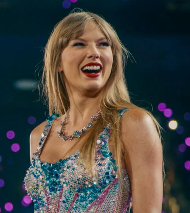 Taylor Swift steht auf der Bühne und trägt ein glitzerndes, buntes Kleid und eine auffällige Halskette vor unscharfem Hintergrund mit lila Lichtpunkten.