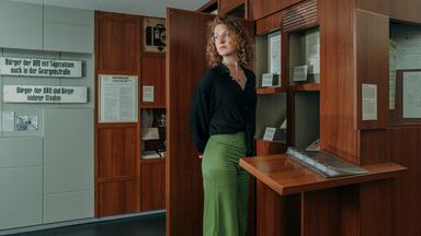 Siska, eine Frau mit lockigen, roten Haaren und Brille, steht in einem Ausstellungsraum im Museum. Ihr Blick geht zur Seite und hinter dem Rücken hat sie die Arme verschränkt. Hinter ihr sieht man zwei Schilder aus DDR Zeiten.
