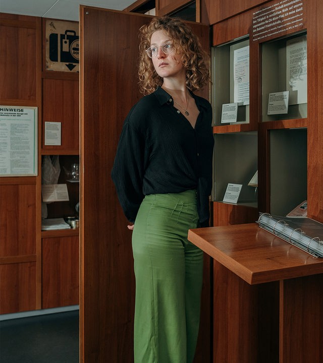 Siska, eine Frau mit lockigen, roten Haaren und Brille, steht in einem Ausstellungsraum im Museum. Ihr Blick geht zur Seite und hinter dem Rücken hat sie die Arme verschränkt. Hinter ihr sieht man zwei Schilder aus DDR Zeiten.
