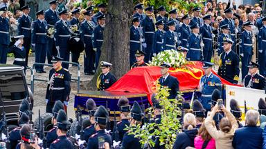 um den Sarg mit rotem Tuch stehen viele Menschen in Uniform