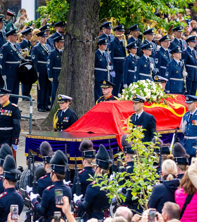 um den Sarg mit rotem Tuch stehen viele Menschen in Uniform
