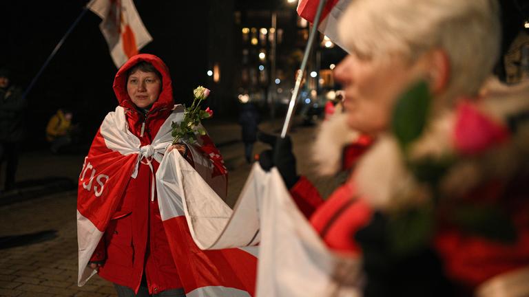 Menschen stehen mit Flaggen und Blumen auf einer Straße.