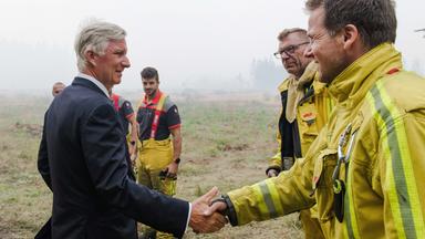 Auf dem Bild ist König Philippe zu sehen. Dieser begrüßt Einsatzkräfte in Feuerwehrschutzkleidung in einem von Rauch betroffenen Naturgebiet. Im Vordergrund schüttelt er einem Feuerwehrmann die Hand, während weitere Einsatzkräfte im Hintergrund stehen. 