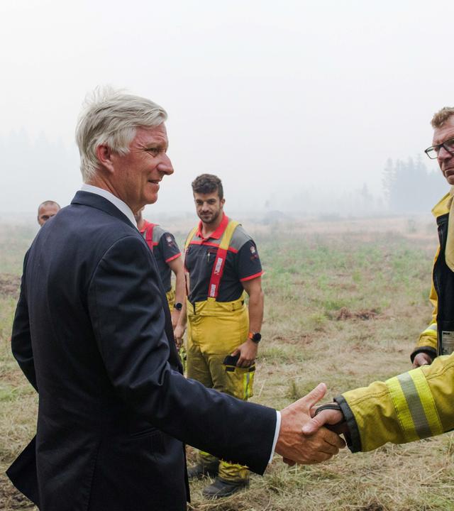 Auf dem Bild ist König Philippe zu sehen. Dieser begrüßt Einsatzkräfte in Feuerwehrschutzkleidung in einem von Rauch betroffenen Naturgebiet. Im Vordergrund schüttelt er einem Feuerwehrmann die Hand, während weitere Einsatzkräfte im Hintergrund stehen. 