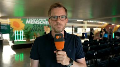 Benthin ist bei der einer Veranstaltung der Partei DIE GRÜNEN zu sehen, hält ein Mikrofon in der Hand un schaut in die Kamera.