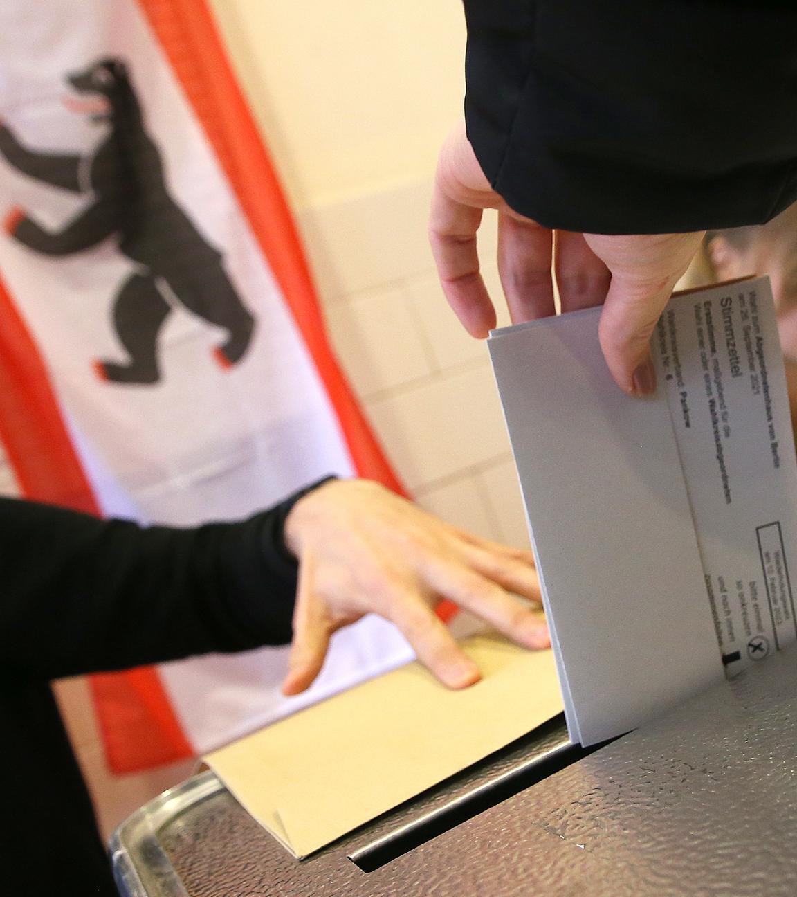 Eine Person wirft ihren Wahlzettel in die Wahlurne. Im Hintergrund sitzen zwei Wahlhelfer an einem Tisch.