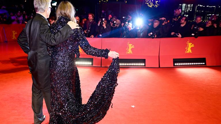 Rückansichteines Mannes im dunklen Smoking und einer Frau im glitzernden Abendkleid mit Schleppe auf dem roten Teppich der Berlinale. Sie wenden sich den Fotografen hinter einer Absperrung zu.