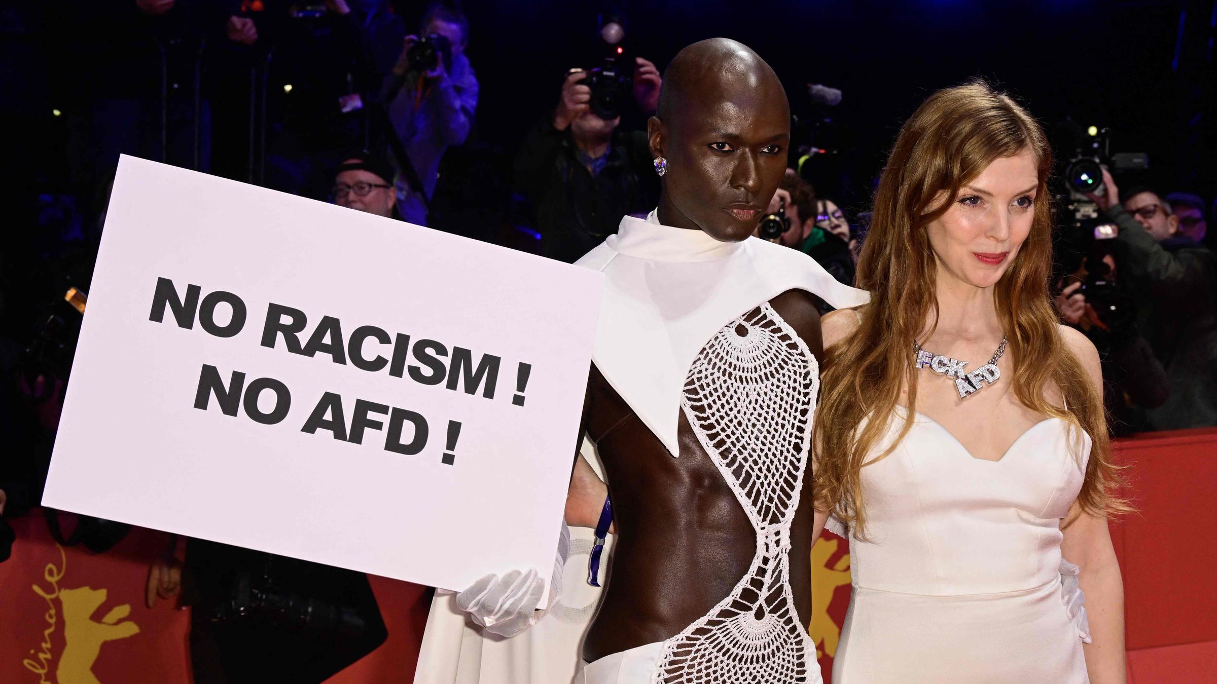 Papis Loveday posiert auf dem roten Teppich mit einem Plakat mit der Aufschrift "No racism no AfD" mit der deutschen Schauspielerin Pheline Roggan, die eine Halskette mit der Aufschrift "FCK AFD" trägt, vor der Eröffnungszeremonie der 74. Berlinale, Europas erstem großen Filmfestival des Jahres, in Berlin am 15.02.2024.
