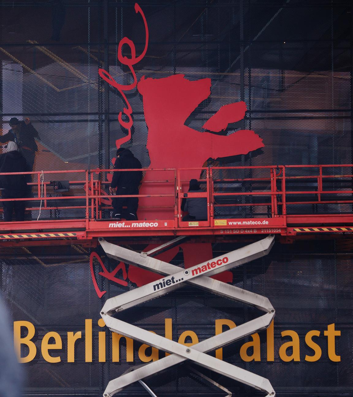 Arbeiter stehen auf einem Podest und installieren an einer Hausfassade das Logo der Berlinale, einen roten Bären.