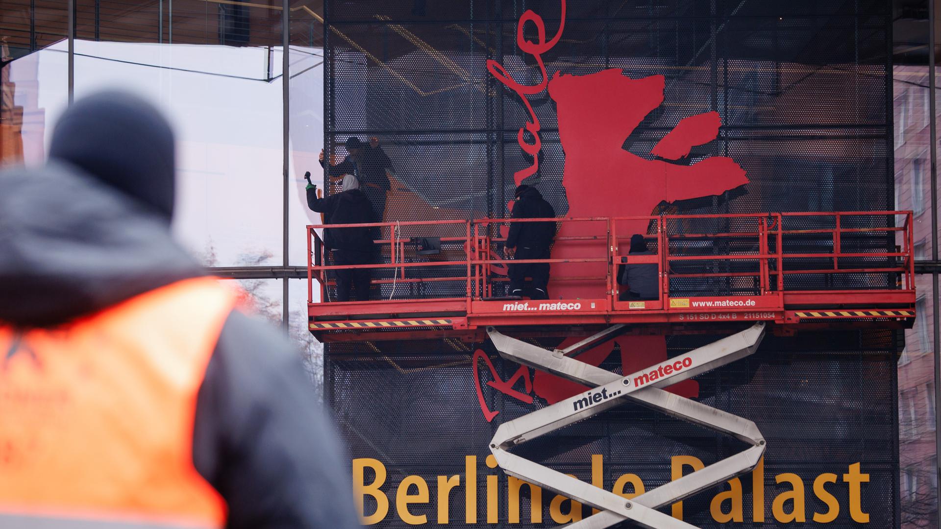 Arbeiter stehen auf einem Podest und installieren an einer Hausfassade das Logo der Berlinale, einen roten Bären.