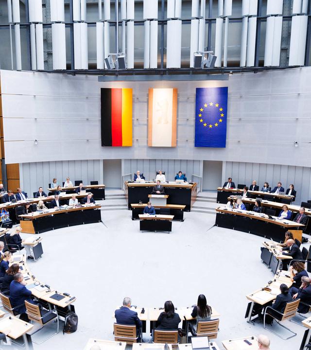 Der Plenarraum des Berliner Abgeordnetenhaus während einer Plenarsitzung. An der Wand hängen drei Flaggen: Deutschland, das Land Berlin und die EU.