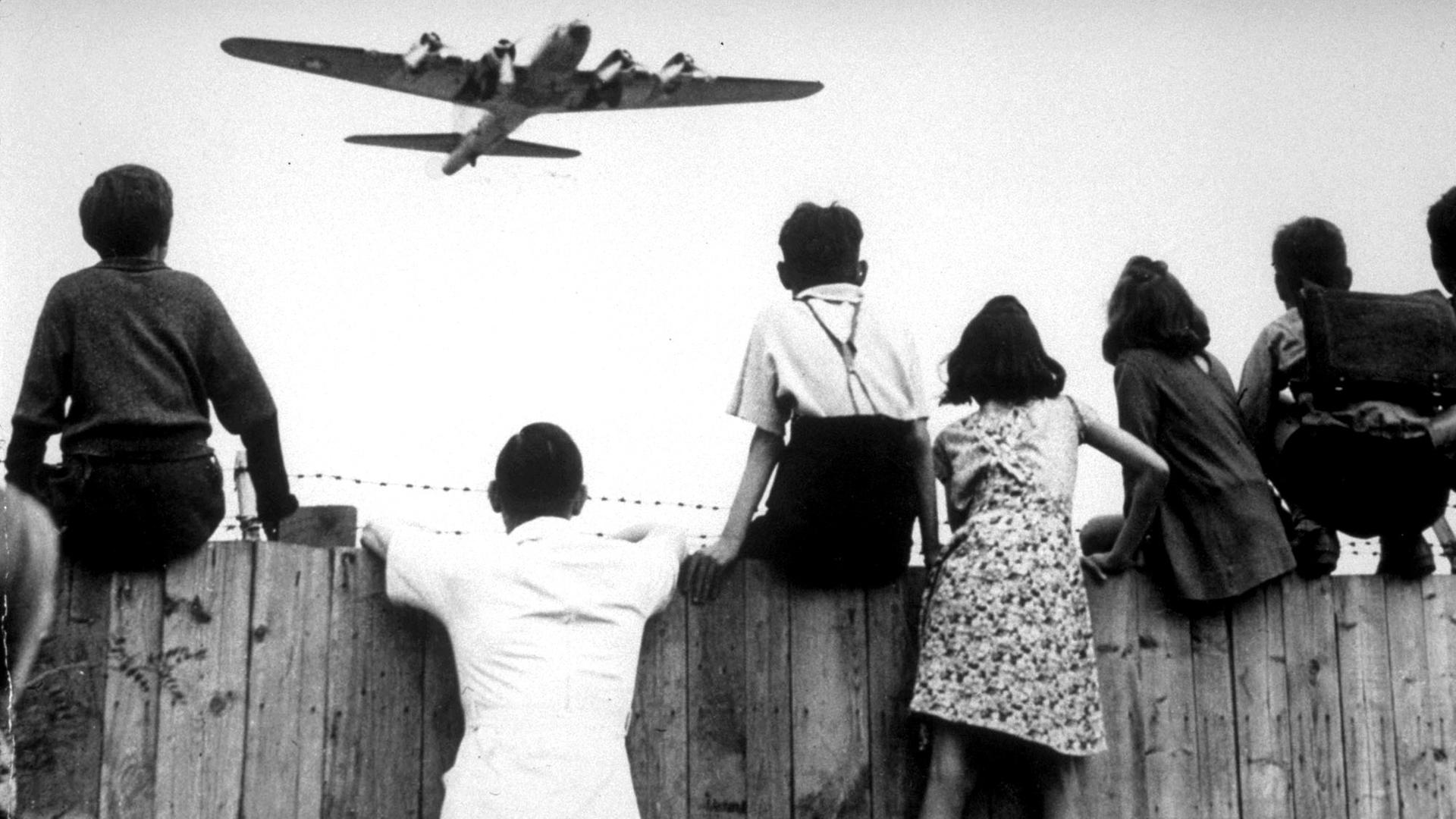 Berliner Kinder sitzen 1948 auf einem Zaun des Berliner Flughafens Tempelhof, während ein Bomber der US-Luftstreitkräfte zur Landung ansetzt
