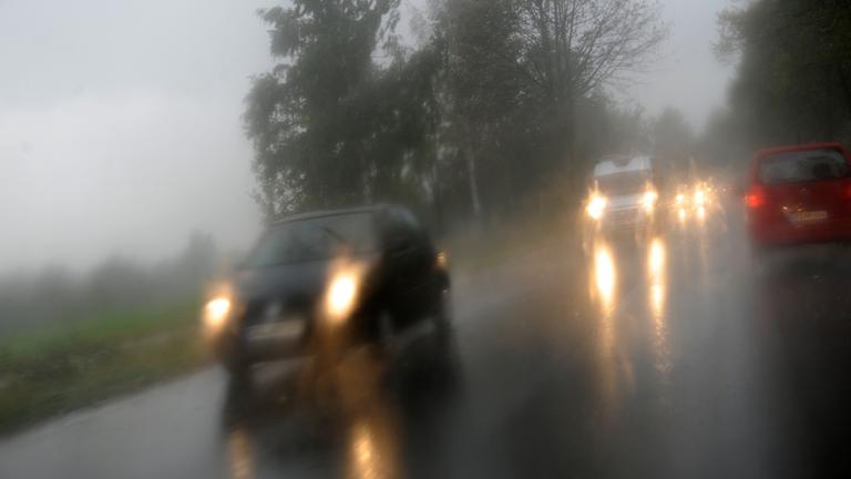 Blick durch die Windschutzscheibe auf schemenhafte Fahrzeuge auf einer Straße während es regnet.