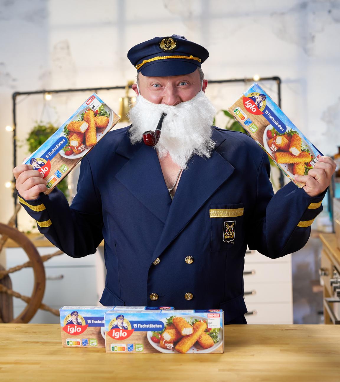 Sebastian Lege steht als Captain Iglo verkleidet und mit Pfeife im Mund vor einem Tisch mit mehreren Packungen Iglo Fischstäbchen.