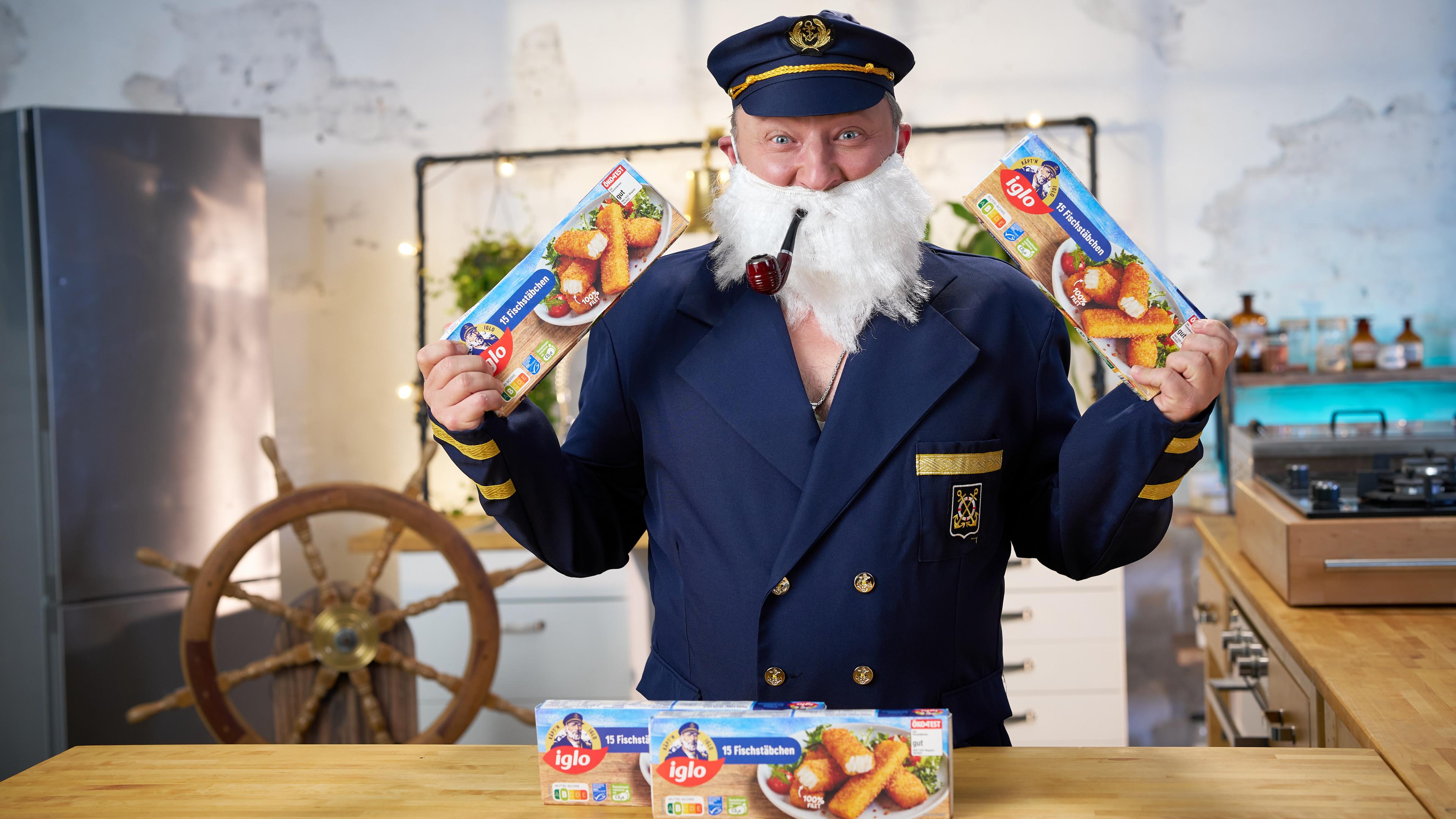 Sebastian Lege steht als Captain Iglo verkleidet und mit Pfeife im Mund vor einem Tisch mit mehreren Packungen Iglo Fischstäbchen.