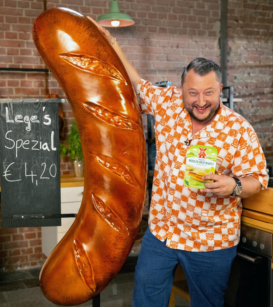 Sebastian Lege steht an eine übergroße Wurst-Reklame gelehnt und hält eine Packung eines pflanzlichen Rügenwalder-Mühle-Produktes in der Hand.