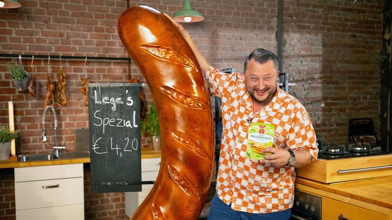 Sebastian Lege steht an eine übergroße Wurst-Reklame gelehnt und hält eine Packung eines pflanzlichen Rügenwalder-Mühle-Produktes in der Hand.
