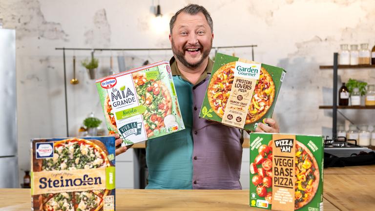 Sebastian Lege lacht frontal in die Kamera. Auf Brusthöhe hält er in jeder Hand einen Pizzakarton. Vor ihm stehen zwei weitere Kartons.