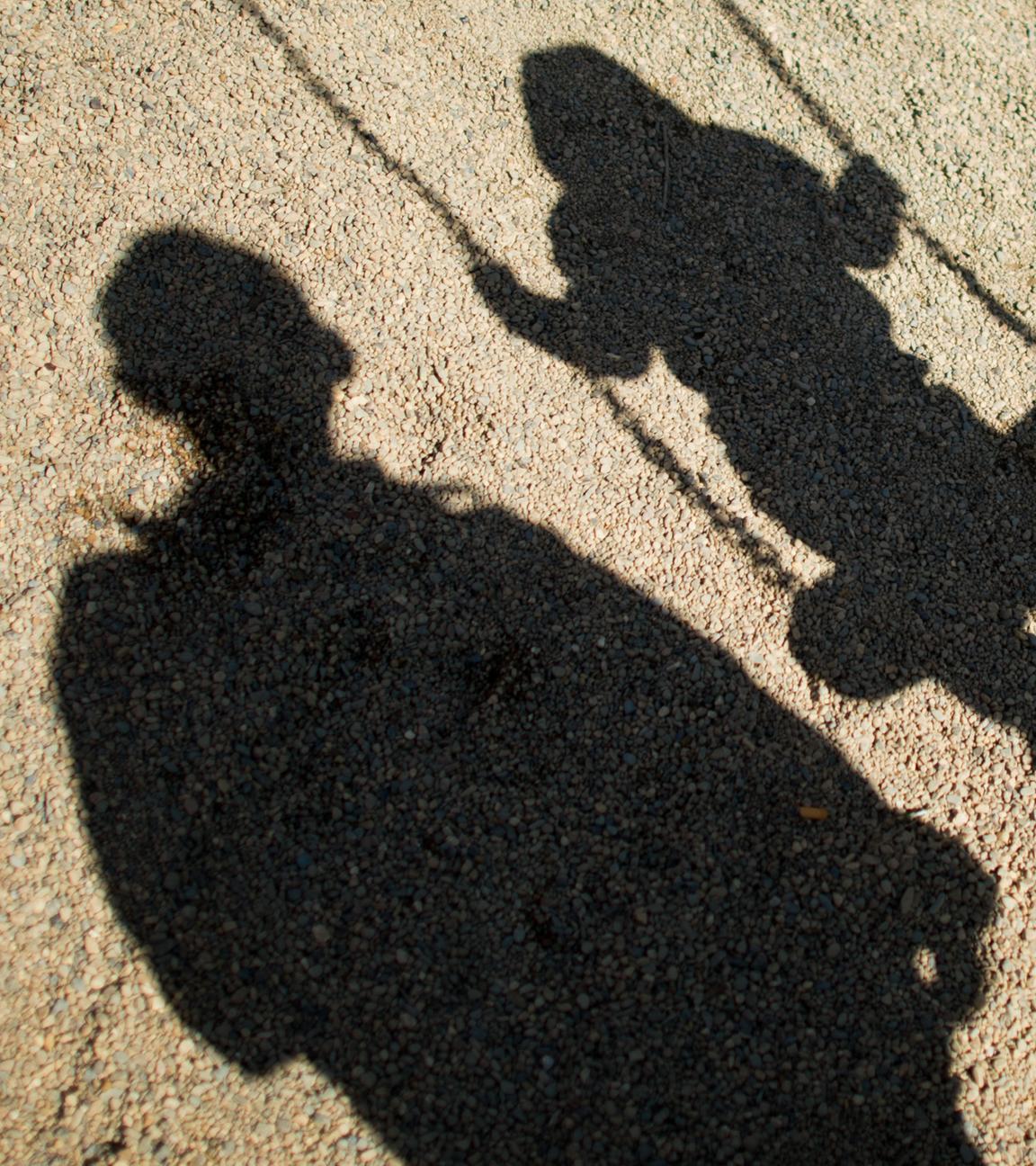 Der Schatten von einem Mann und einem schaukelnden Kind fallen auf Sand auf einem Spielplatz.