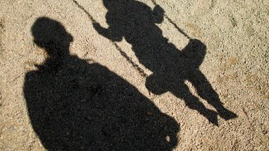 Der Schatten von einem Mann und einem schaukelnden Kind fallen auf Sand auf einem Spielplatz.