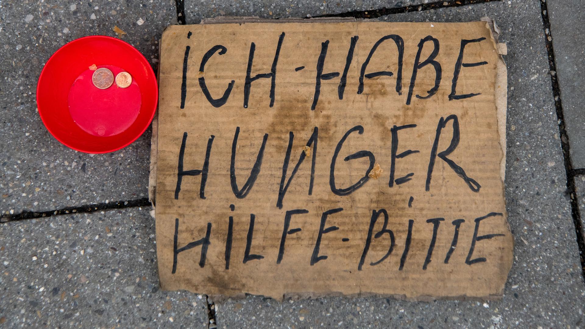 Eine rote Schale mit ein paar Cent-Geldstücken und ein Pappschild mit der Aufschrift "ich habe Hunger - Hilfe bitte"