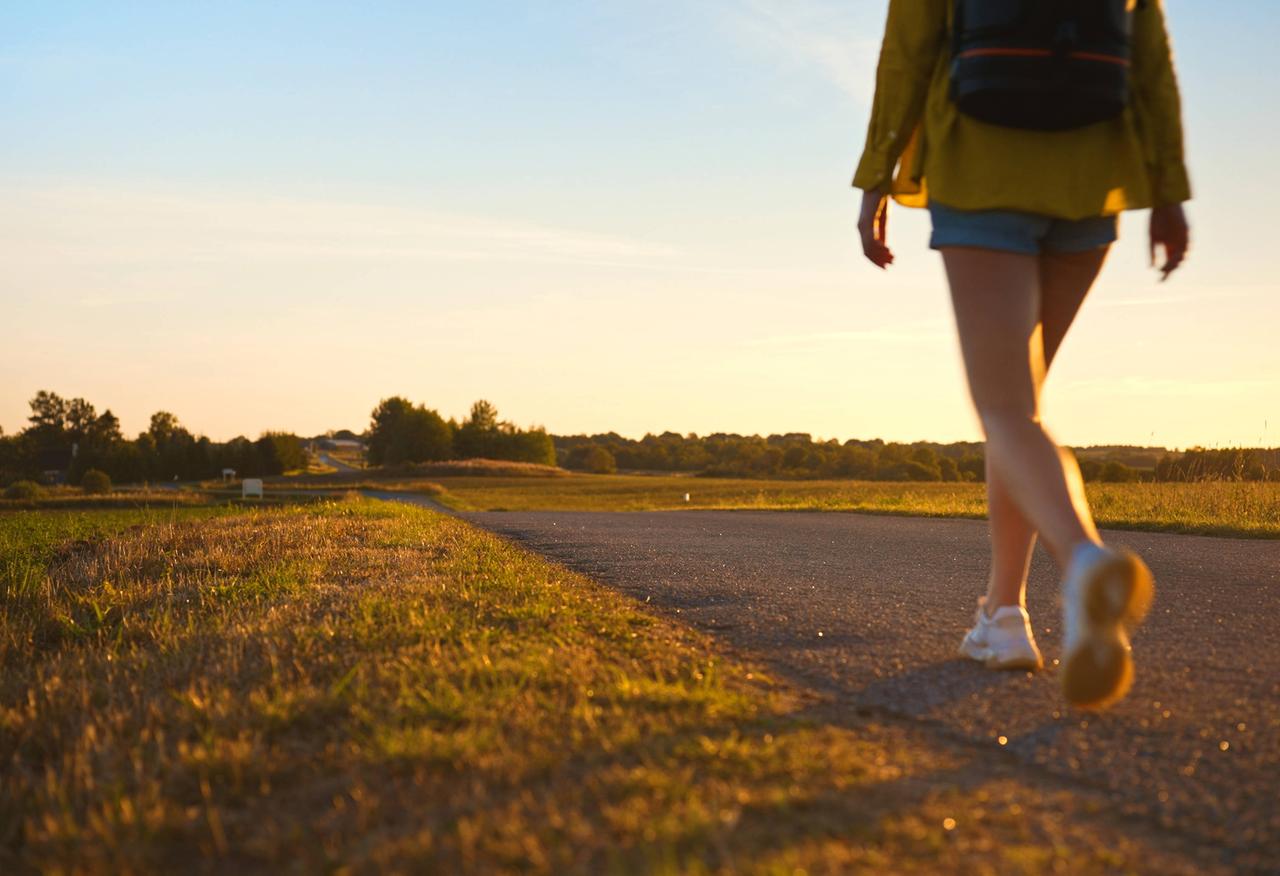 Symbolbild: Frau mit Rucksack geht entlang der Straße in den Sonnenuntergang.