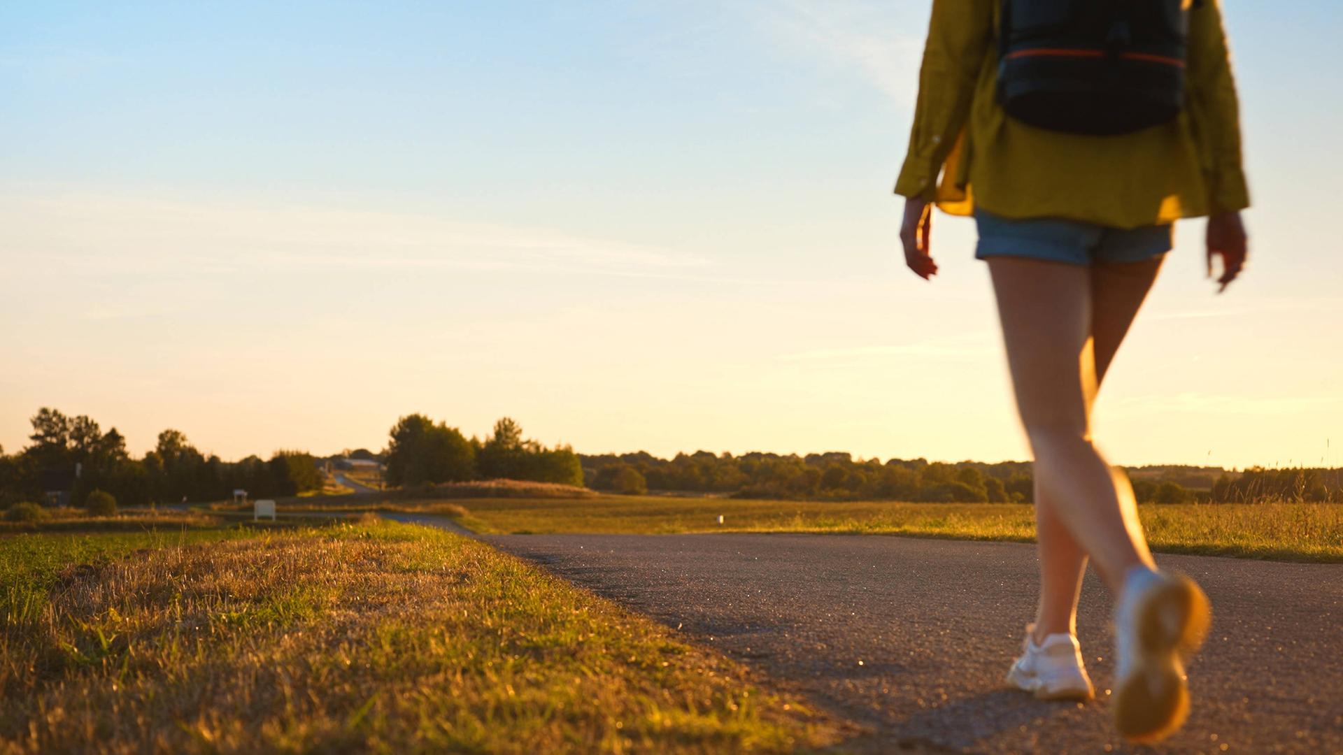 Symbolbild: Frau mit Rucksack geht entlang der Straße in den Sonnenuntergang.