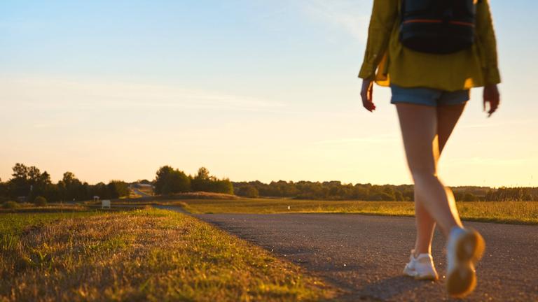 Symbolbild: Frau mit Rucksack geht entlang der Straße in den Sonnenuntergang.