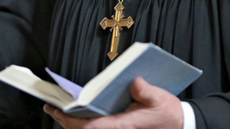 Ein Pfarrer mit Kreuzkette und schwarzem Gewand hält eine aufgeschlagene Bibel in seinen Händen.