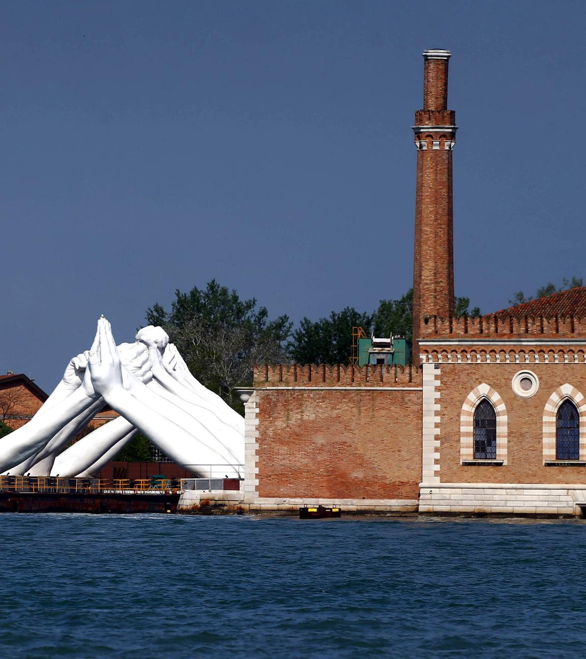 Neben einem alten Ziegelsteingebäude mit Turm am Wasser ist eine große weiße Skulptur zu sehen, die aus Händen besteht, die sie paarweise an der Hand anfassen.