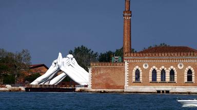 Neben einem alten Ziegelsteingebäude mit Turm am Wasser ist eine große weiße Skulptur zu sehen, die aus Händen besteht, die sie paarweise an der Hand anfassen.