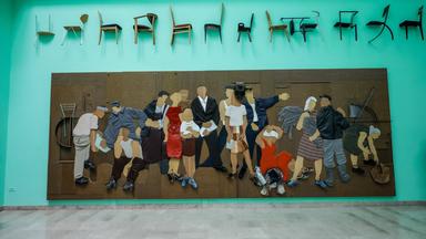 An einer mintgrünen Wand hängt ein großes Kunstwerk, das eine große Gruppe Menschen zeigt. Darüber sind in einer Reihe Stühle an der Wand befestigt.