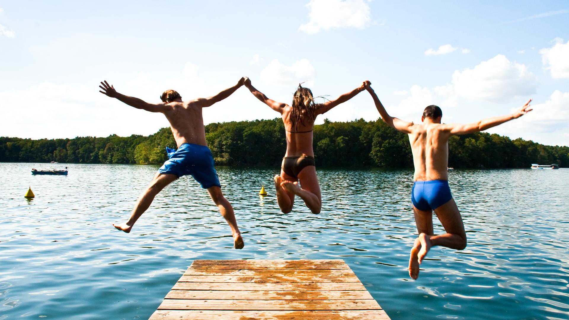 Zwei Jungs und ein Mädchen springen von einem Steg ins Wasser. Sie halten sich dabei an den Händen.