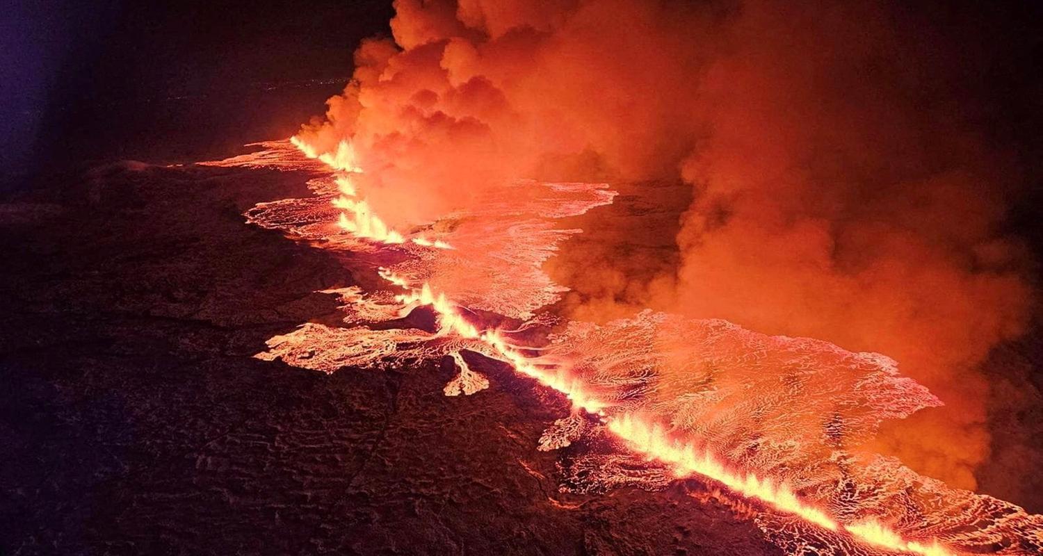Eine Luftaufnahme über einer Eislandschaft in der dunklen Nacht zeigt mitten drin einen Vulkanausbruch. Ein langer glühender spalt spuckt Feuer und Rauch, während sich kleine verästelte, leuchtende Flüsse von ihm ausgehend wie Adern ausbreiten.