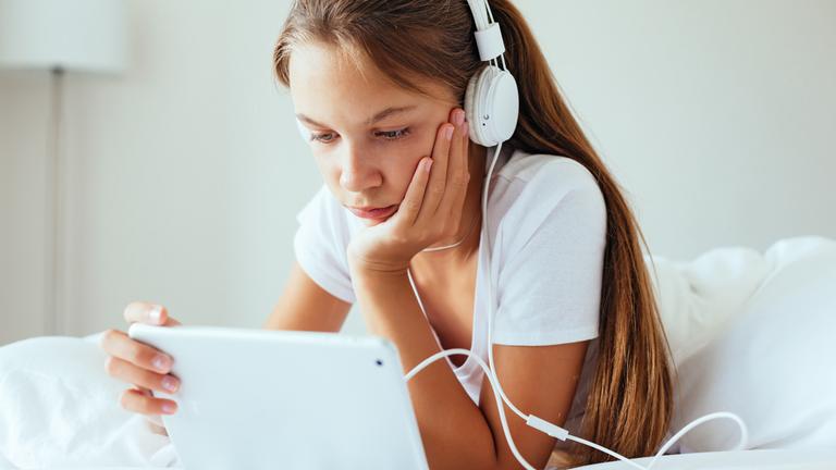 Ein Mädchen liegt auf ihrem Bett. Sie trägt Kopfhörer und schaut auf ein Tablet.