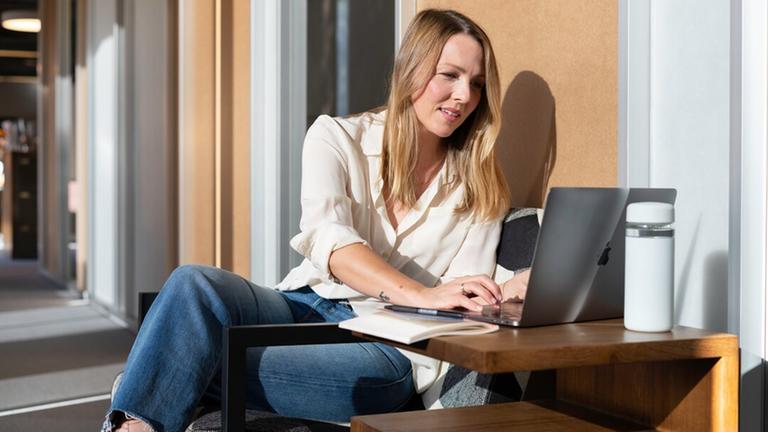 Eine Frau sitzt auf einem Balkon in der Sonne. Sie schaut lachend auf einen Laptop.