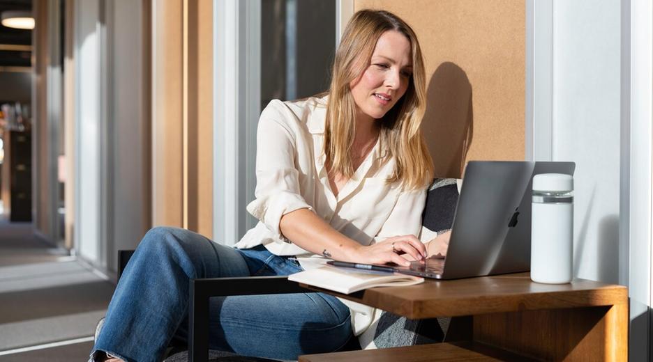 Eine Frau sitzt auf einem Balkon in der Sonne. Sie schaut lachend auf einen Laptop.