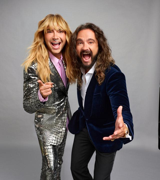 Bill und Tom Kaulitz Bill und Tom Kaulitz stehen vor einem grauen Hintergrund und beugen sich zur Kamera. Bill trägt einen glänzenden Anzug, Tom ein dunkles Jackett. Seitlich sind Studiowände und Stative zu sehen.