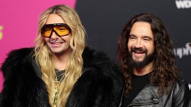 Auf dem Bild sind zu sehen: Bill Kaulitz steht links neben Tom Kaulitz. Beide sind mit Kopf und Oberkörper zu sehen und lächeln. 