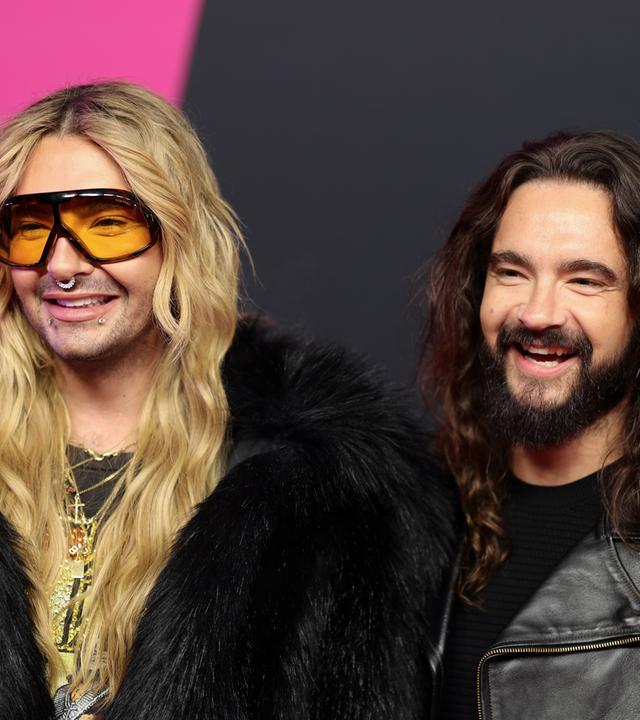 Auf dem Bild sind zu sehen: Bill Kaulitz steht links neben Tom Kaulitz. Beide sind mit Kopf und Oberkörper zu sehen und lächeln. 