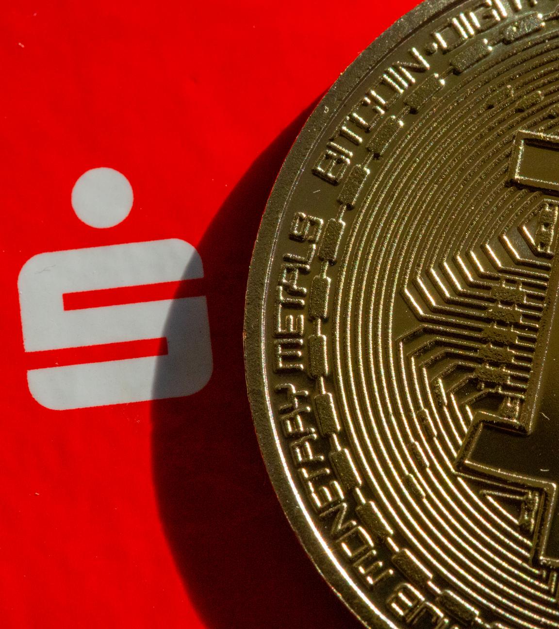 Eine Bitcoin-Münze liegt neben dem Logo der Sparkasse.
