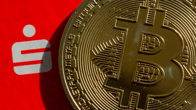 Eine Bitcoin-Münze liegt neben dem Logo der Sparkasse.