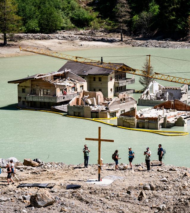 Bewohner des Lötschentals stehen an einem See mit Häusern und einem Kreuz, das die Stelle der zerstörten Kirche markiert, während einer Gedenkfeier ein Jahr nach dem Erdrutsch von Blatten.