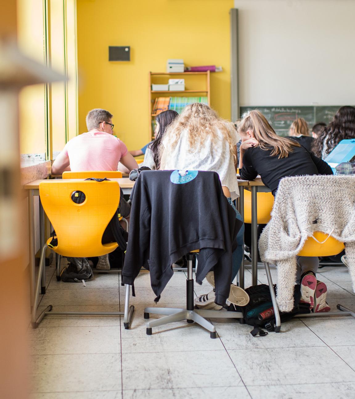 Schüler eines Gymnasiusms sitzen während des Unterrichts im Klassenzimmer. Sie werden von hinten fotografiert, sodass man sie sitzend auf den Stühlen sieht, wie sie ihre Köpfe zusammenstecken.