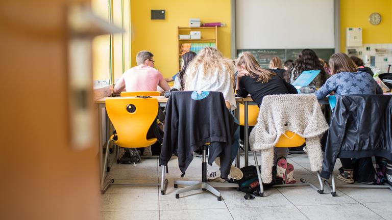 Schüler eines Gymnasiusms sitzen während des Unterrichts im Klassenzimmer. Sie werden von hinten fotografiert, sodass man sie sitzend auf den Stühlen sieht, wie sie ihre Köpfe zusammenstecken.