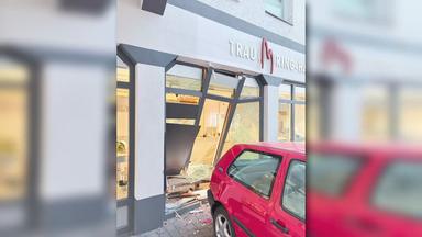 Rotes Auto vor einem Geschäft mit beschädigter Schaufensterfront. Mehrere Fensterscheiben sind zerbrochen, Teile der Fassade und Glassplitter liegen vor dem Gebäude.