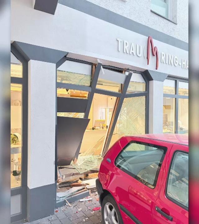 Rotes Auto vor einem Geschäft mit beschädigter Schaufensterfront. Mehrere Fensterscheiben sind zerbrochen, Teile der Fassade und Glassplitter liegen vor dem Gebäude.
