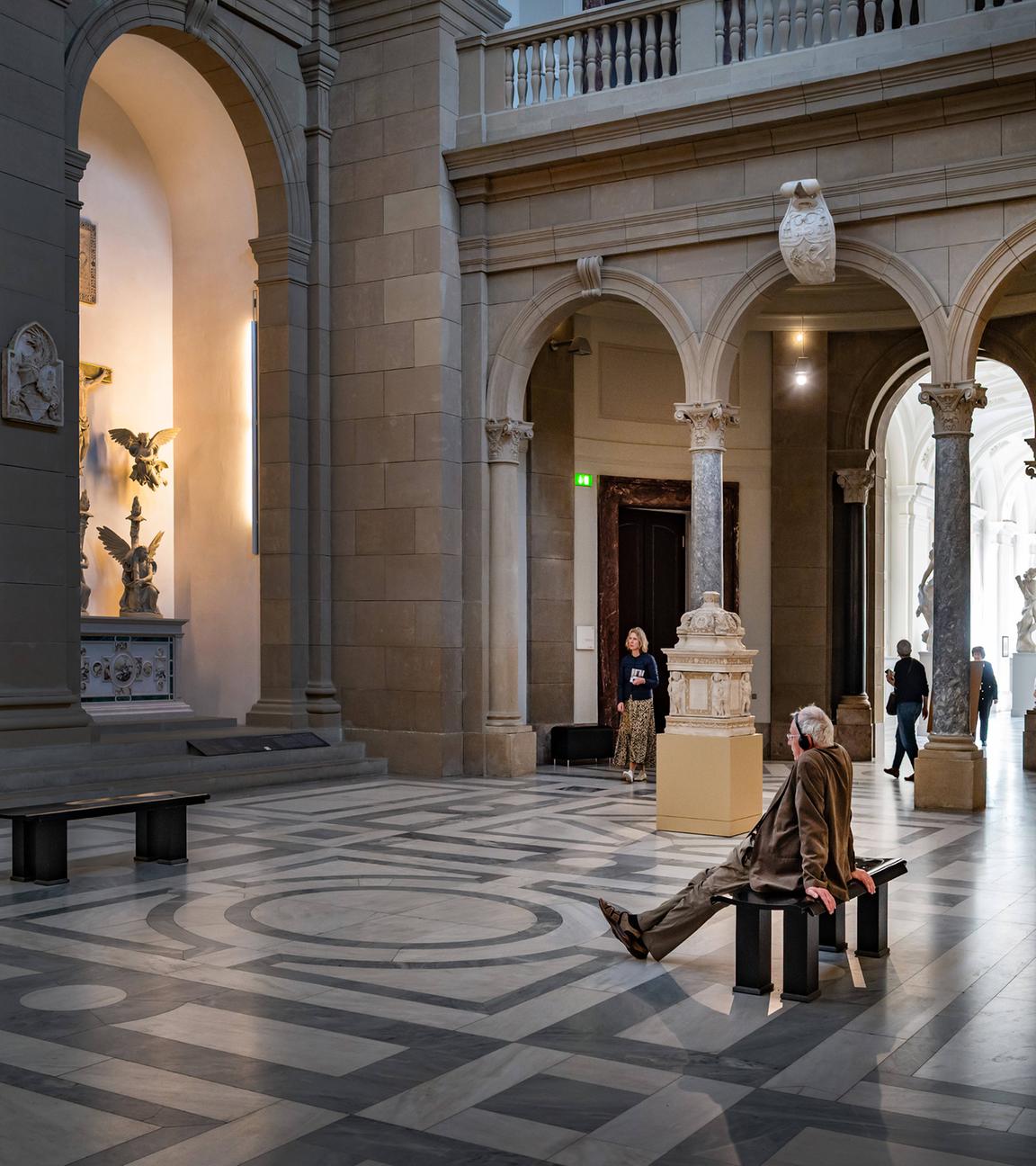 Besicher sitzt in Museumraum auf einer Bank und betrachtet Kunstwerke.